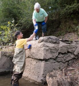 A man in waders standing in the river handing a bag full of water to a woman above him on the shore.